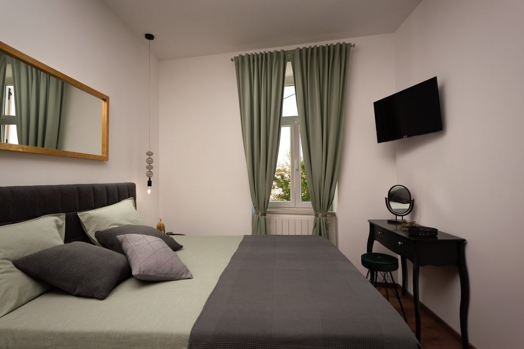 A bedroom with a bed, a desk, a chair, a mirror, a TV, and a window with curtains.