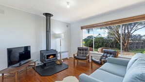 A modern living room with a wood-burning stove, a flat-screen TV, leather furniture, and a large window with a view of trees. - Willan - 43 Powling Street (Port Fairy)