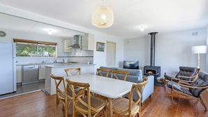 A modern kitchen with white cabinets, a dining area with wooden chairs, a sofa, and a fireplace. - Willan - 43 Powling Street (Port Fairy)