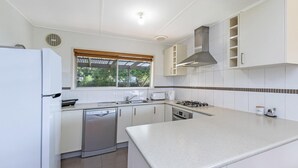 A compact kitchen with white cabinets, a built-in dishwasher, a gas stove, and a window with blinds. - Willan - 43 Powling Street (Port Fairy)