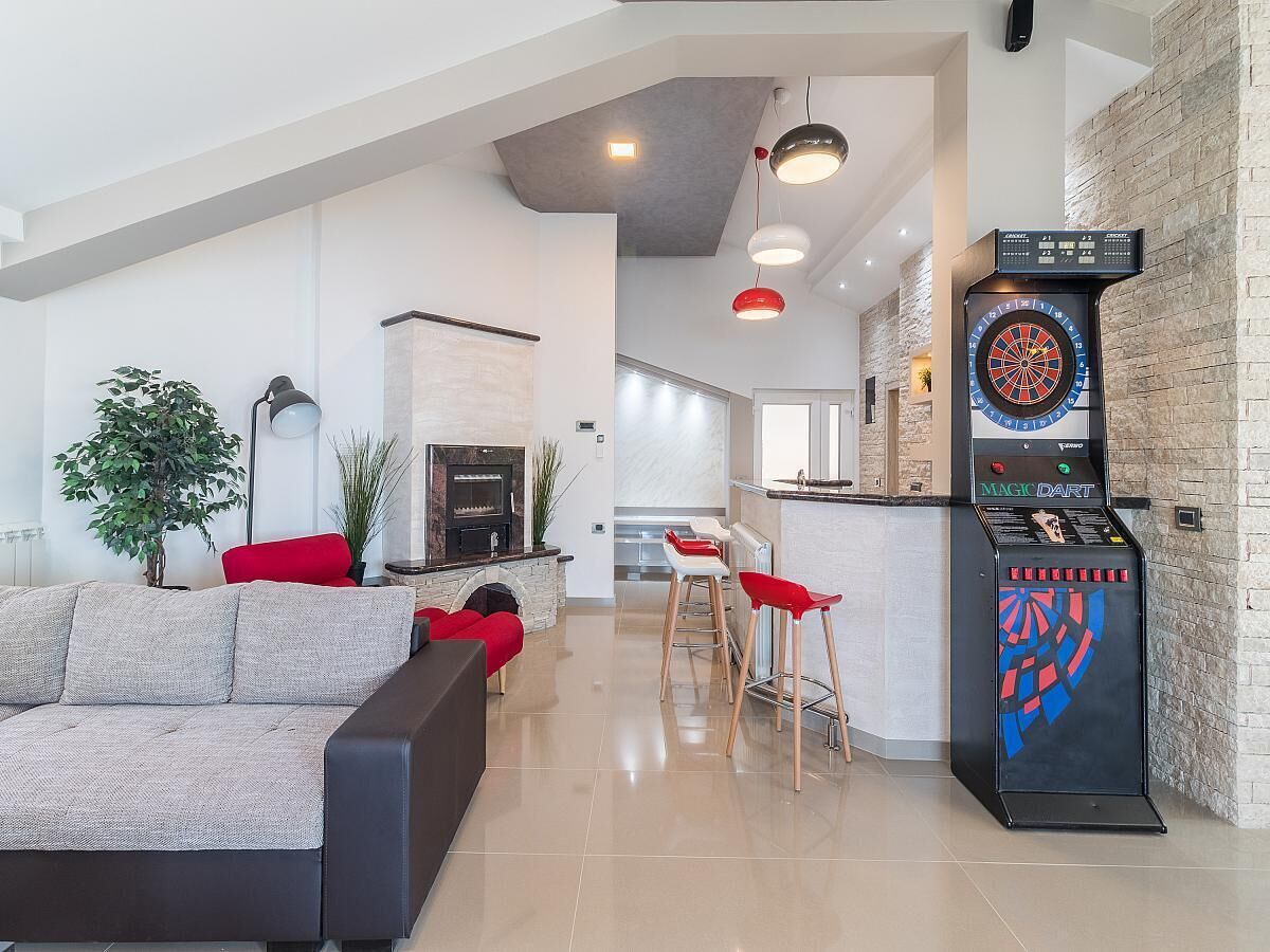 A modern living room with a fireplace, a dartboard, and a kitchen area in the background.