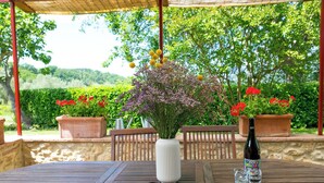 En udendørs terrasse med et træbord, en hvid vase med blomster og plantekasser.
