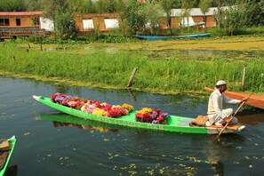 A green boat with colorful flowers and the text "Mr Marvelous Flower Man" on a calm water body with greenery and buildings in the background. - Golden Crest Houseboats (Srinagar)