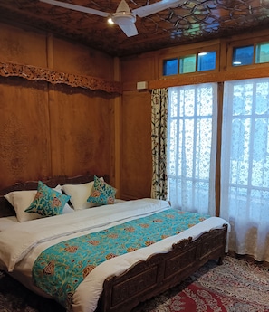 Quarto de madeira com cama, um ventilador de teto e uma janela com cortinas florais.