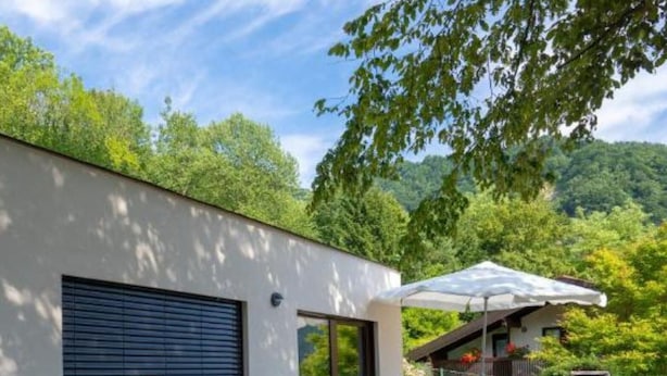 A modern building with a stone retaining wall, a covered outdoor seating area, and a view of a forested hillside. - Nature View House With Sauna (Tolmin)