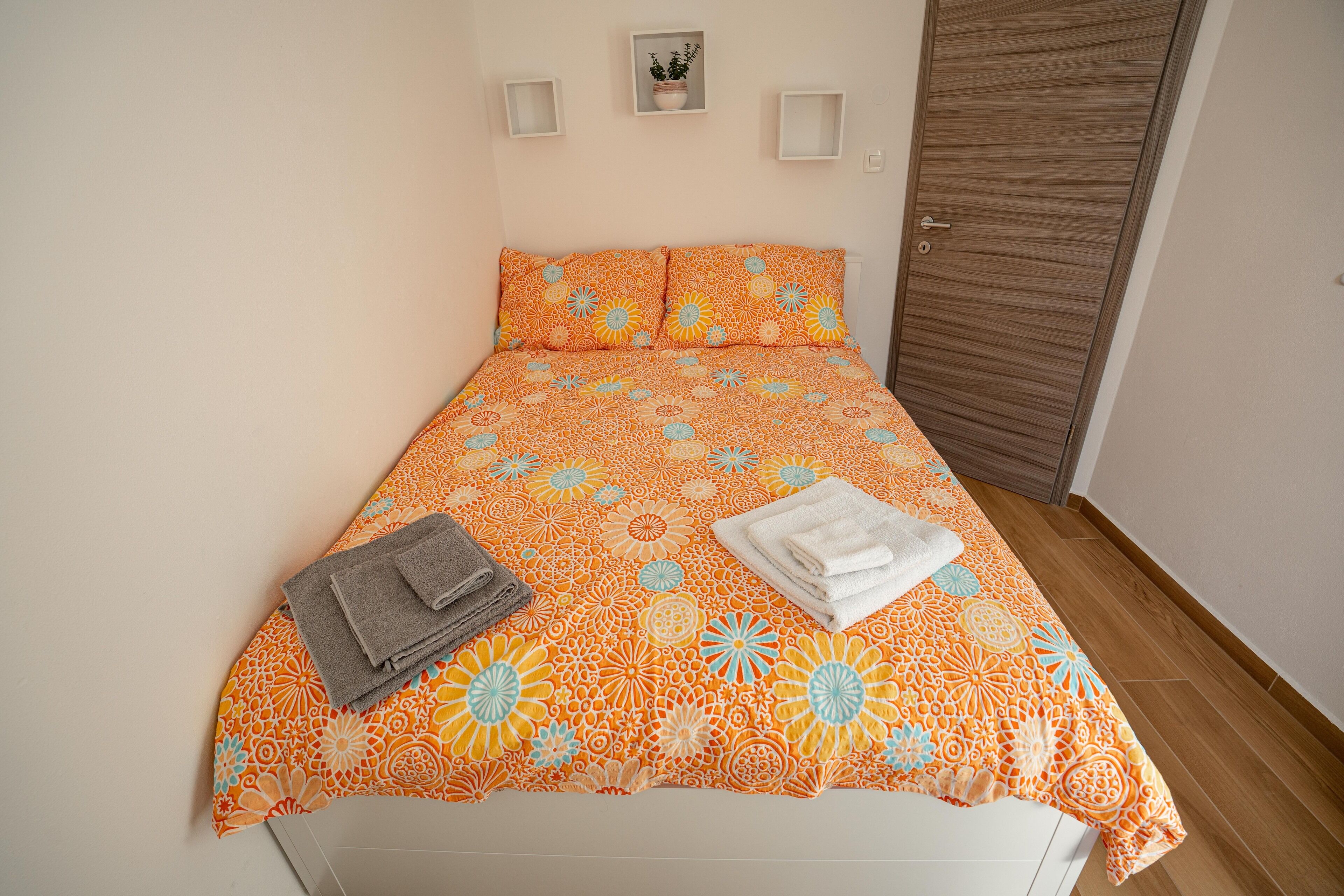 A bed with an orange floral bedspread, two neatly folded towels, and a small potted plant on the wall.