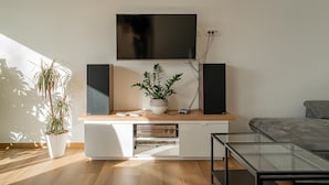 A modern living room with a flat-screen TV mounted on the wall, a wooden entertainment unit with speakers, a potted plant, and a glass coffee table. - Nature View House With Sauna (Tolmin)