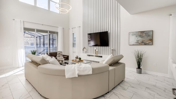 A modern living room with a curved sectional sofa, a flat-screen TV mounted on the wall, and a large window providing natural light. - 5Bd Home, Storey Lake with Private Pool 14 Guests - Private Pool - 2773 BD (Kissimmee)