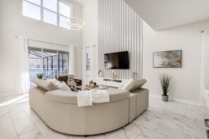 Un salon moderne doté d’un grand canapé d’angle, d’un téléviseur à écran plat fixé au mur et d’une grande fenêtre laissant entrer la lumière naturelle.