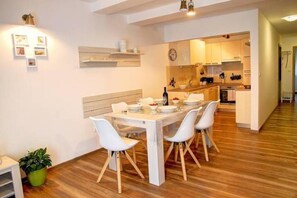 Un comedor con una mesa blanca dispuesta para cuatro personas, rodeada de sillas blancas. Se observa un suelo de madera y una planta en maceta.