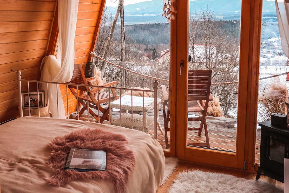 A cozy wooden cabin bedroom with a large bed, a view through a glass door to a snowy landscape, and a rustic interior.