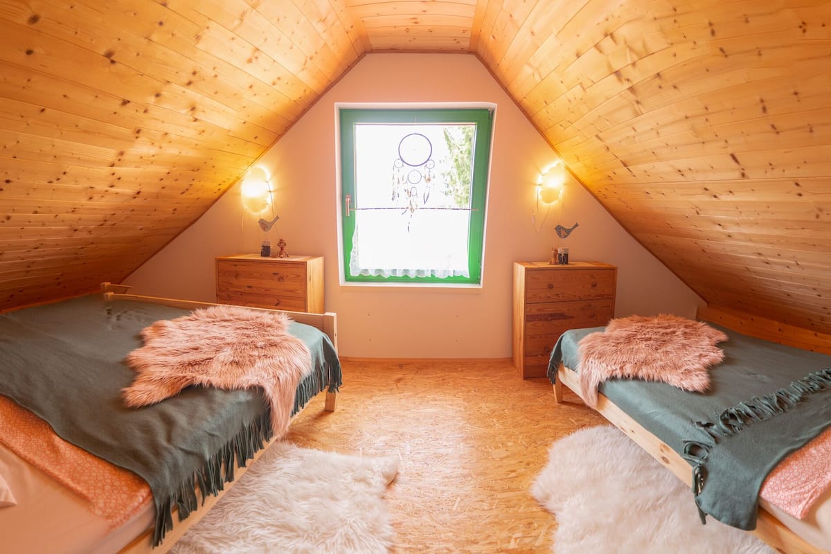 A cozy attic bedroom with two beds, wooden walls, a window with a view, and a small dresser.