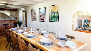 A dining area with a long wooden table set for a meal, framed artwork on the wall, and a view into the kitchen. - Villa écrin charance (Gap)