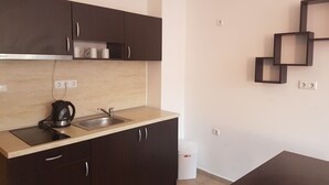 Une cuisine moderne avec des meubles de couleur sombre, un évier en acier inoxydable et une bouilloire. On y trouve une poubelle blanche et un luminaire fixé au mur.