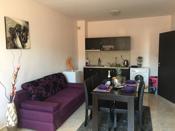 A compact living space with a purple sofa, a dining table set for two, a kitchen area with a washing machine, and a decorative piece on the wall. - Salt Lake Aparthotel (Pomorie)