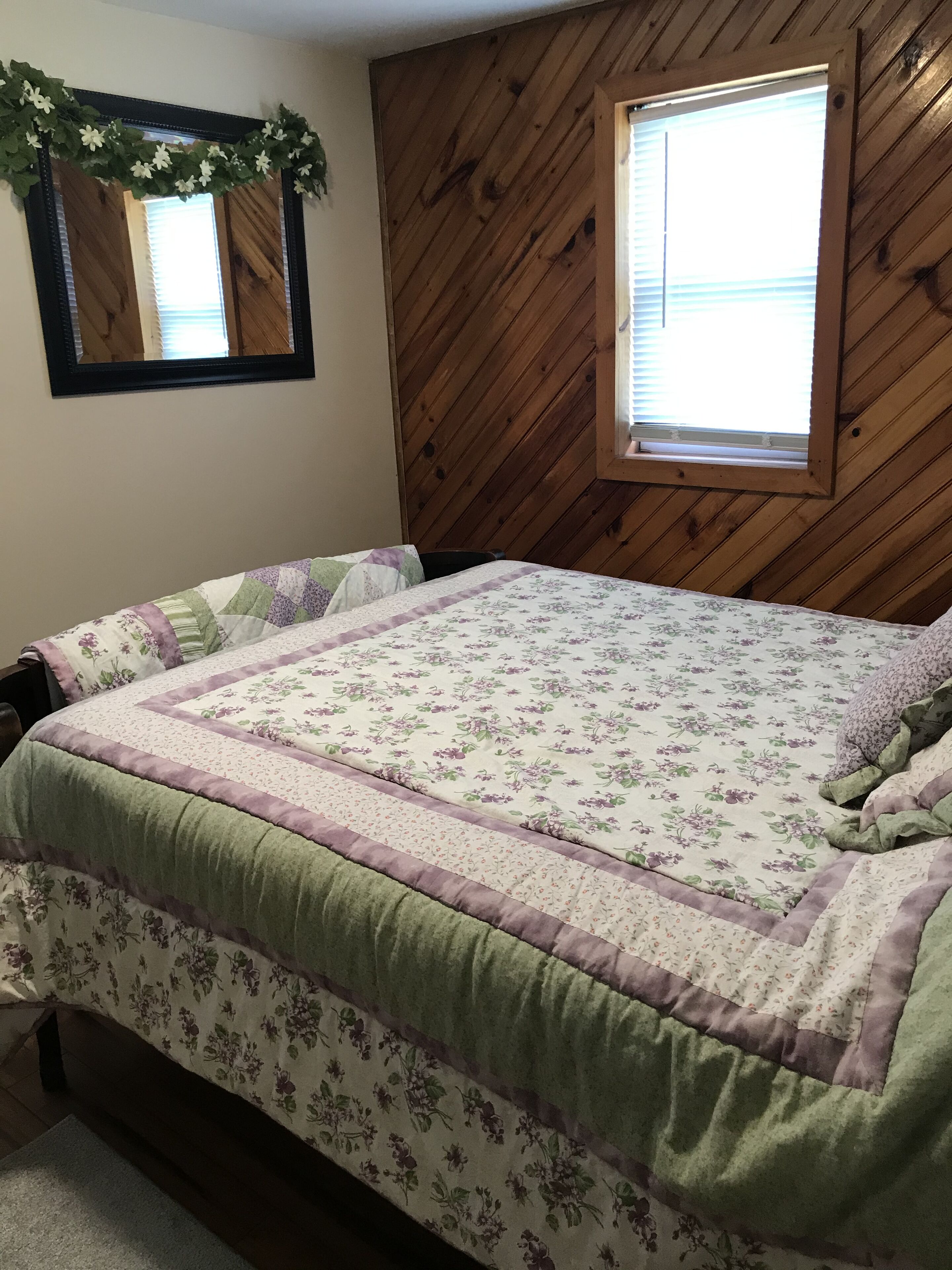 Ein Schlafzimmer mit holzverkleideter Schräge, einem Bett mit floraler Bettwäsche, einem Fenster mit Jalousien und einem Spiegel mit Blumenkranz.