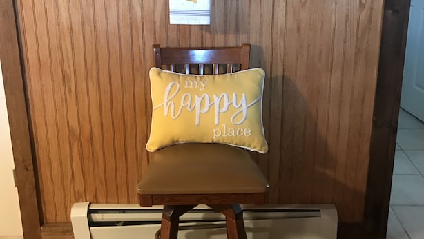 A wooden wall with framed pictures and a potted plant. A chair with a cushion labeled "my happy place" is in front.