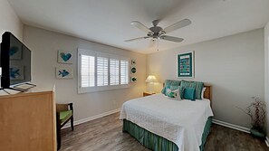 Une chambre à coucher comprenant un lit, un ventilateur de plafond, une fenêtre avec des stores et un bureau avec une chaise.