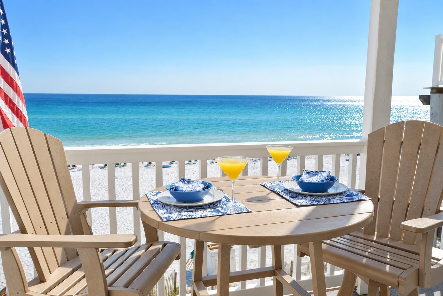 A wooden deck with two chairs and a table, overlooking a beach with a clear view of the ocean.