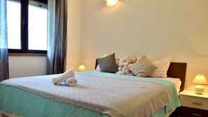 A neatly made bed with a rolled towel, a teal bedspread, and multiple pillows. There are two bedside lamps and a nightstand with a drawer.