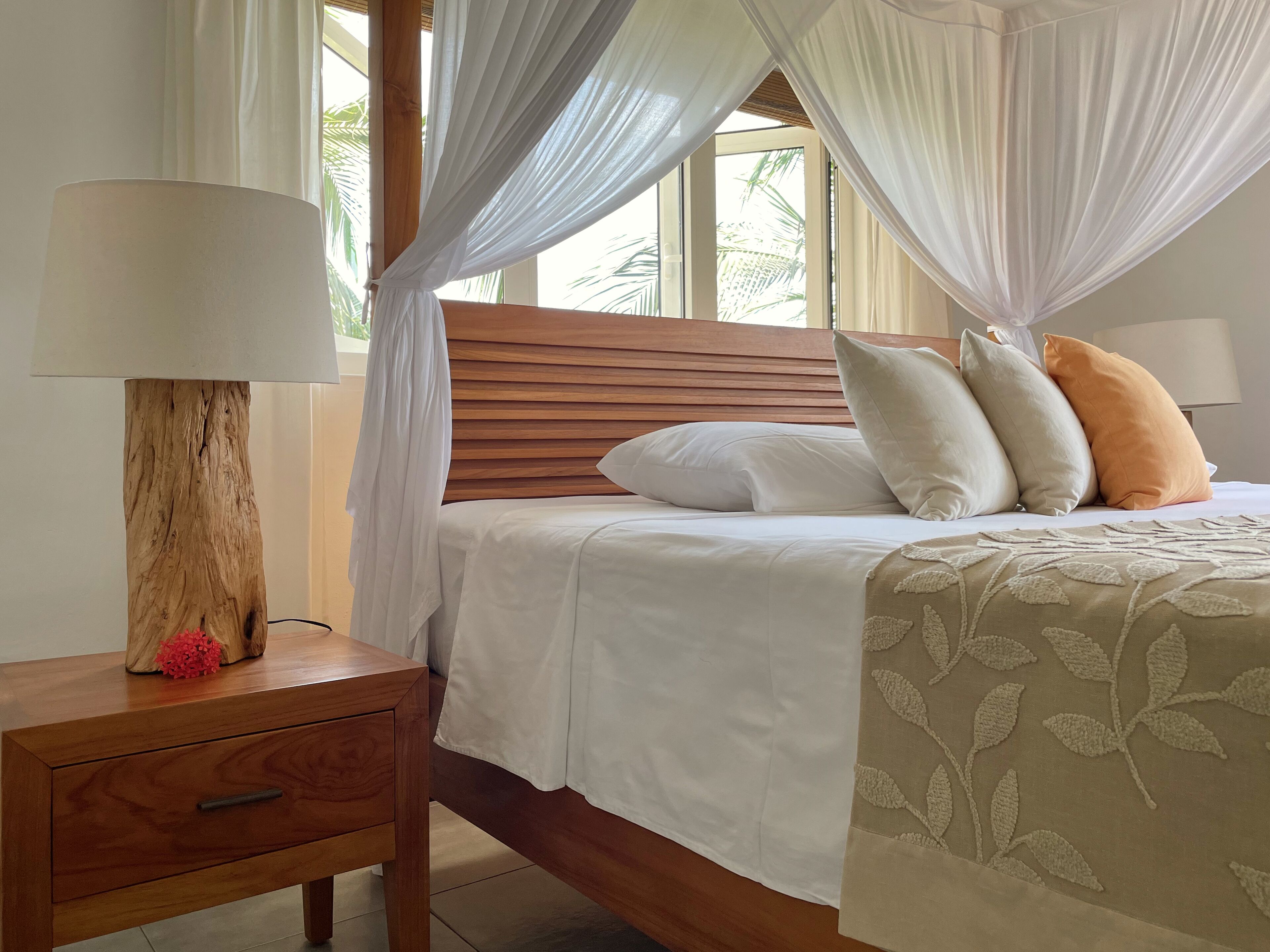A bedroom with a wooden headboard, a bed with white bedding and multiple pillows, a bedside table with a lamp and a flower, and a window with sheer curtains.