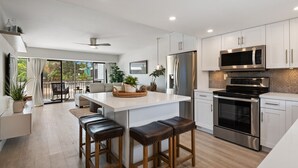 A modern kitchen with white cabinets, a central island with stools, stainless steel appliances, and a sliding glass door leading to a balcony. - Key Largo Villa, Kawama J3, 3/3, Ocean,Marina,Lagoon,Pools,Kayaks&bikes (Key Largo)