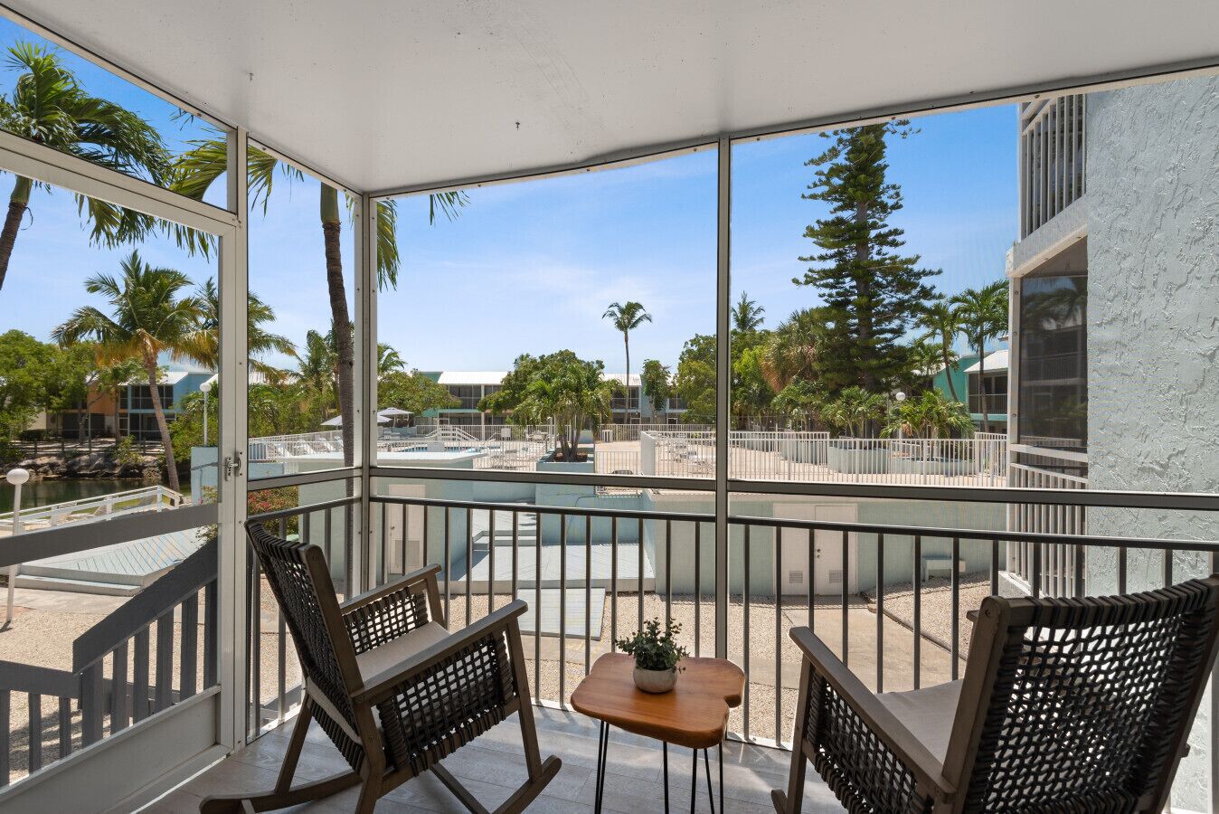 Un balcón con dos sillas de mimbre y una mesita pequeña, con vista a una zona de piscina con palmeras y edificios al fondo.
