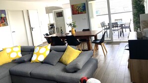 A modern living room with a grey sofa, yellow and grey cushions, a wooden dining table, and a large window offering a view of the outside.