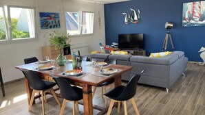 A modern living room with a dining area, featuring a wooden dining table, black chairs, a grey sofa, and a flat-screen TV.