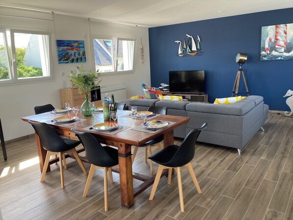 Un salon moderne avec un coin repas, comprenant une table à manger en bois, des chaises noires, un canapé gris et un téléviseur à écran plat.