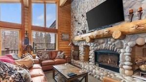 A log cabin interior with a stone fireplace, leather sofas, a television mounted on a wooden beam, and a large window. - Abode at Black Bear #403, hot tub, pool & gym, steps to trails (Park City)
