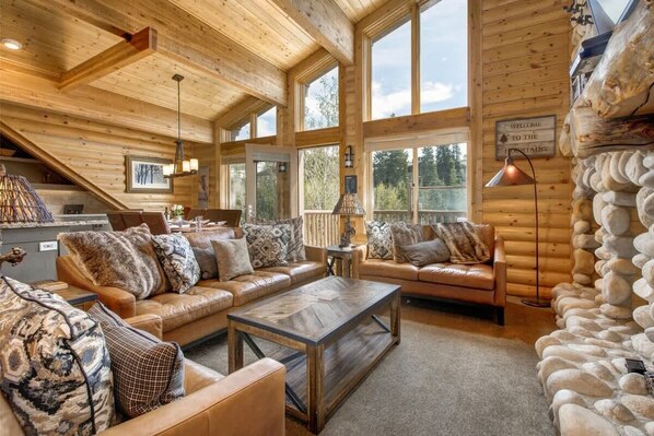 A log cabin interior with a wooden ceiling, large windows, a fireplace, and a stone wall.
