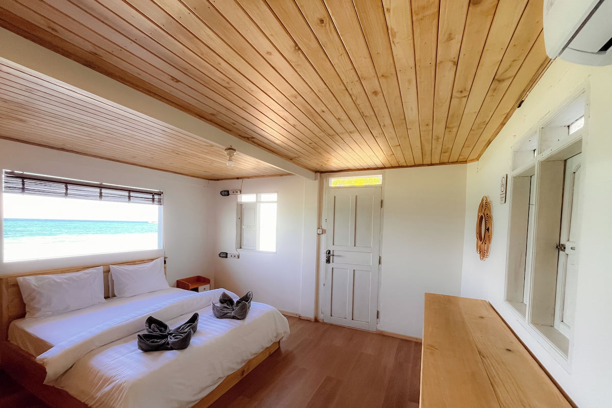 Un dormitorio con techo de madera, una cama con ropa de cama blanca, una mesita de noche pequeña y una ventana con vista al mar.
