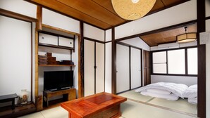 Ein traditioneller japanischer Raum mit Tatami-Böden, einem Fernseher auf einem Holzständer, einem Holztisch und einem Bett mit weißer Bettwäsche.
