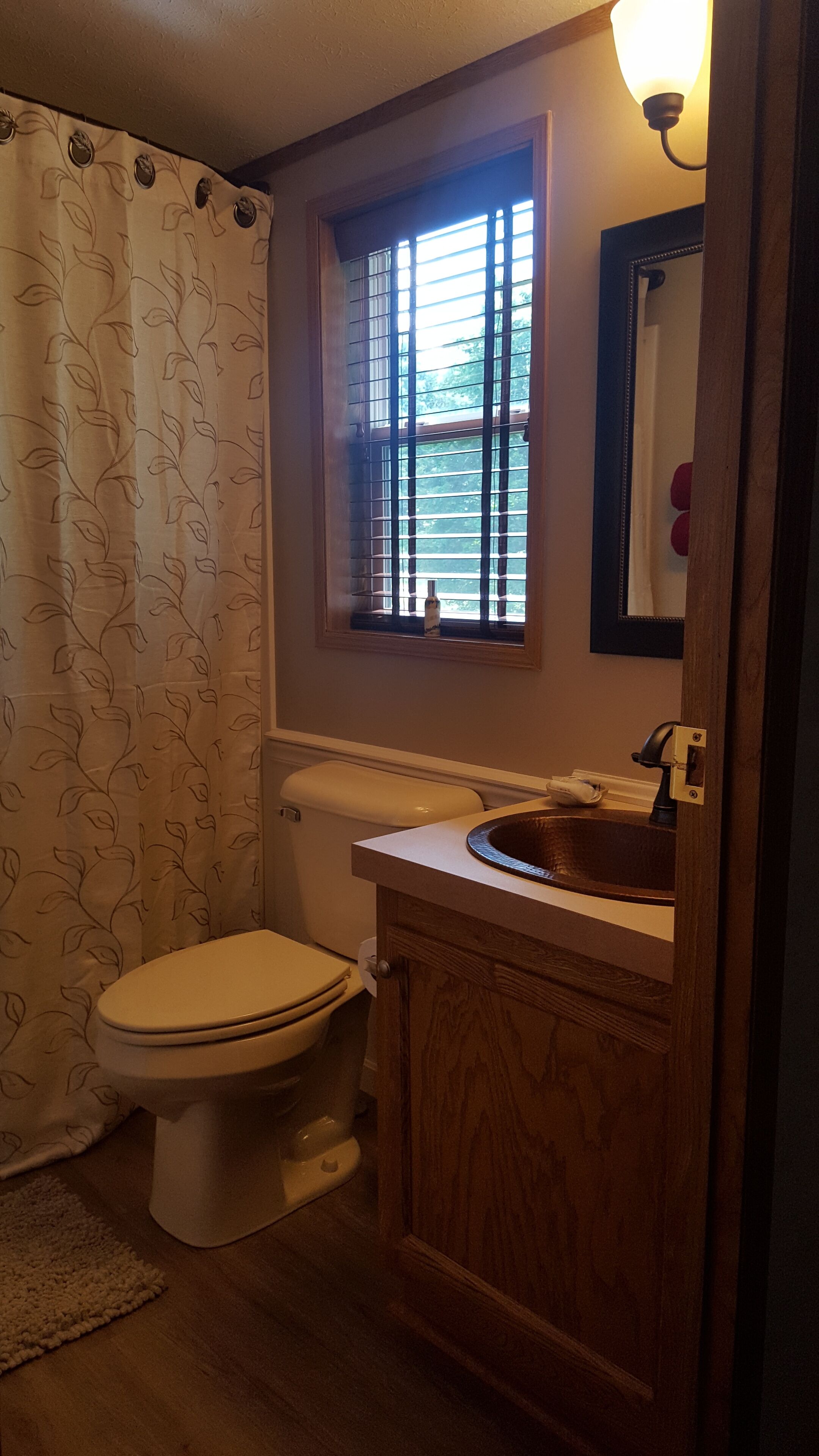 A bathroom with a toilet, a wooden vanity, a window with blinds, and a shower curtain with a leaf pattern.