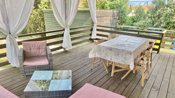 A covered outdoor seating area with wicker furniture and a table, surrounded by a wooden deck and a view of greenery and buildings. - Sunny island (Stari Grad)