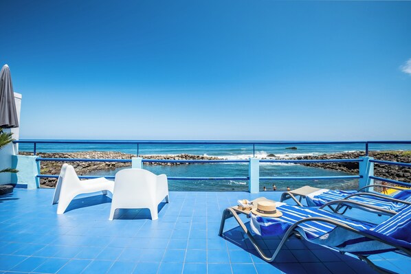 A terrace with blue flooring, white chairs, and striped lounge chairs facing a rocky coastline.