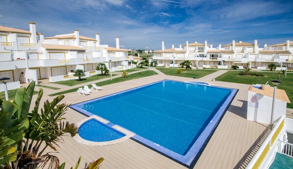 Una piscina grande rodeada por una terraza, con edificios blancos al fondo.
