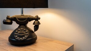 A black rotary telephone on a wooden table with a lamp casting a warm light. - Queen Anne's Home (Great Yarmouth)