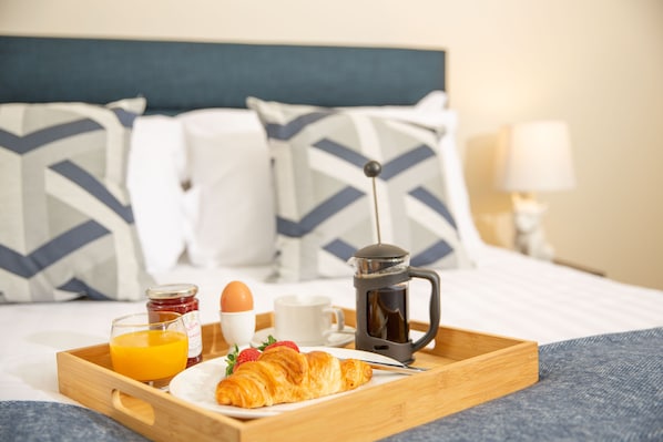 A breakfast tray with croissant, orange juice, jam, and coffee on a bed with a headboard and pillows. - Queen Anne's Home (Great Yarmouth)