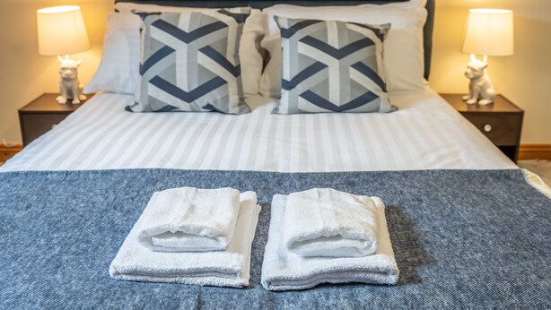 A neatly made bed with striped bedding, two pillows with a chevron pattern, and two folded towels on a blue blanket. - Queen Anne's Home (Great Yarmouth)