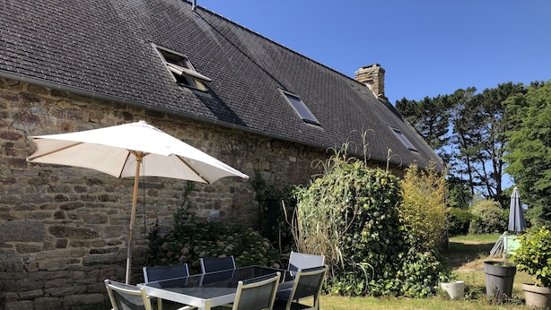 An outdoor patio with a table and chairs, a large umbrella, and a stone building with a tiled roof. - Gite Alcove in a farmhouse beach and coastal path on foot (Névez)