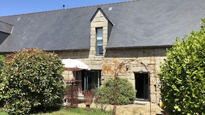 A stone building with a grey roof, a small window, and a door. There is a garden with a wooden table and chairs, and a white umbrella. - Gite Alcove in a farmhouse beach and coastal path on foot (Névez)