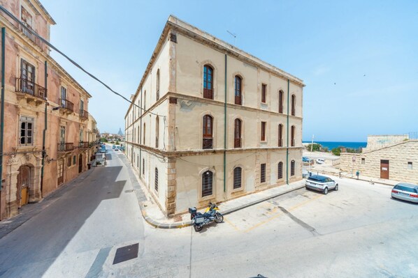 A street with a building featuring a corner entrance, a motorcycle parked in front, and a clear view of the sea in the background.