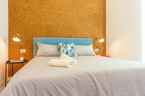Un lit bien fait, avec une tête de lit bleue, du linge de lit blanc et une serviette pliée. Il y a deux tables de chevet, chacune avec une lampe, et un petit vase sur l’une d’elles.