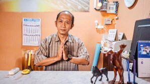 A man seated at a desk with a calendar, a phone, and a computer monitor. - Roomz Budget (Semarang)