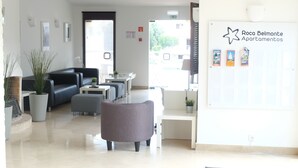 A modern reception area with a black leather sofa, grey armchairs, and a white reception desk. There are potted plants and a bulletin board with property listings. - Roca Belmonte Apartamentos (Albufeira)