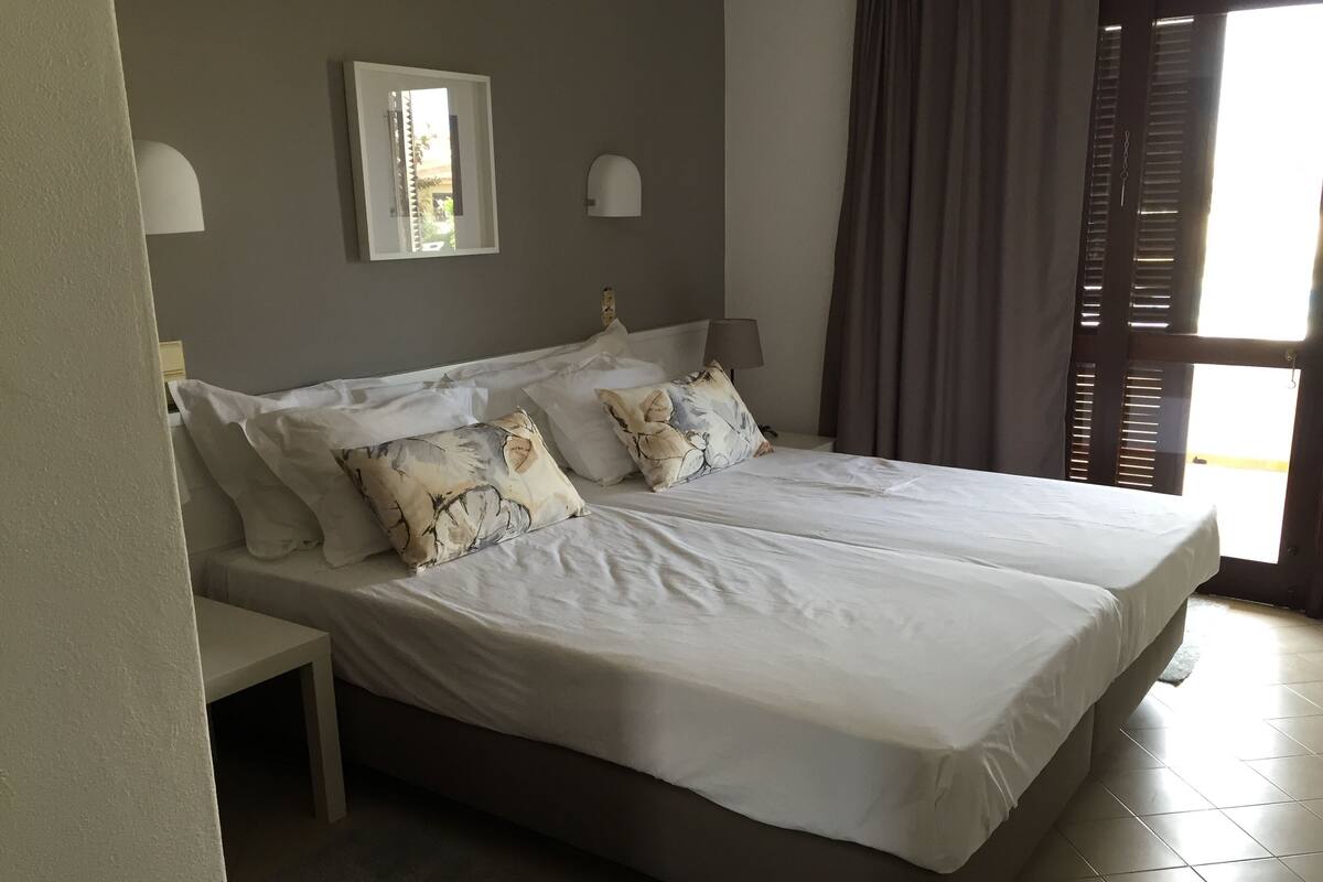 Un lit double avec du linge de lit blanc et des coussins à motifs, une table de chevet, une lampe fixée au mur et une fenêtre avec des rideaux.