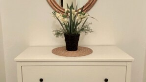 Uma cômoda branca com um espelho redondo acima, um jogo americano trançado e um vaso com flores brancas.