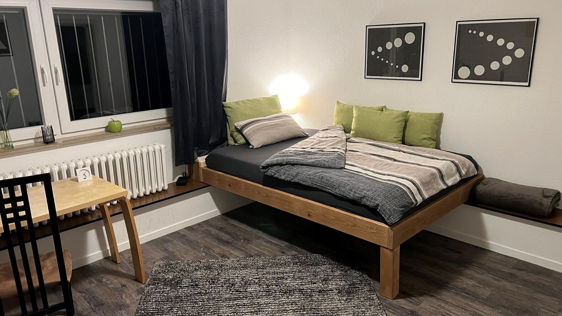 A bedroom with a wooden bed, a desk, a chair, a radiator, and two wall art pieces.
